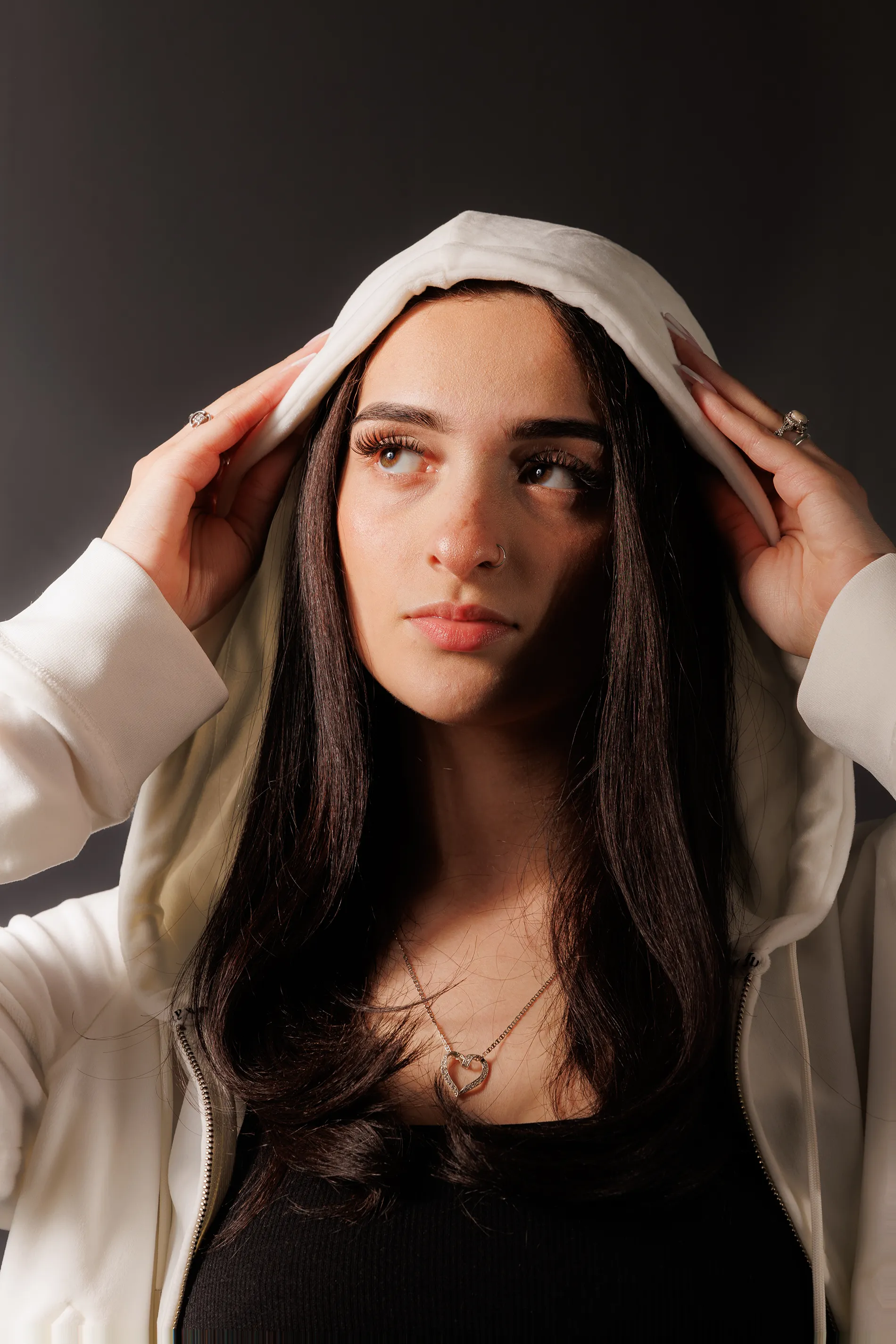 Close-up portrait of Areli wearing a white hoodie against a dark backdrop, shot in Monticello, NY.