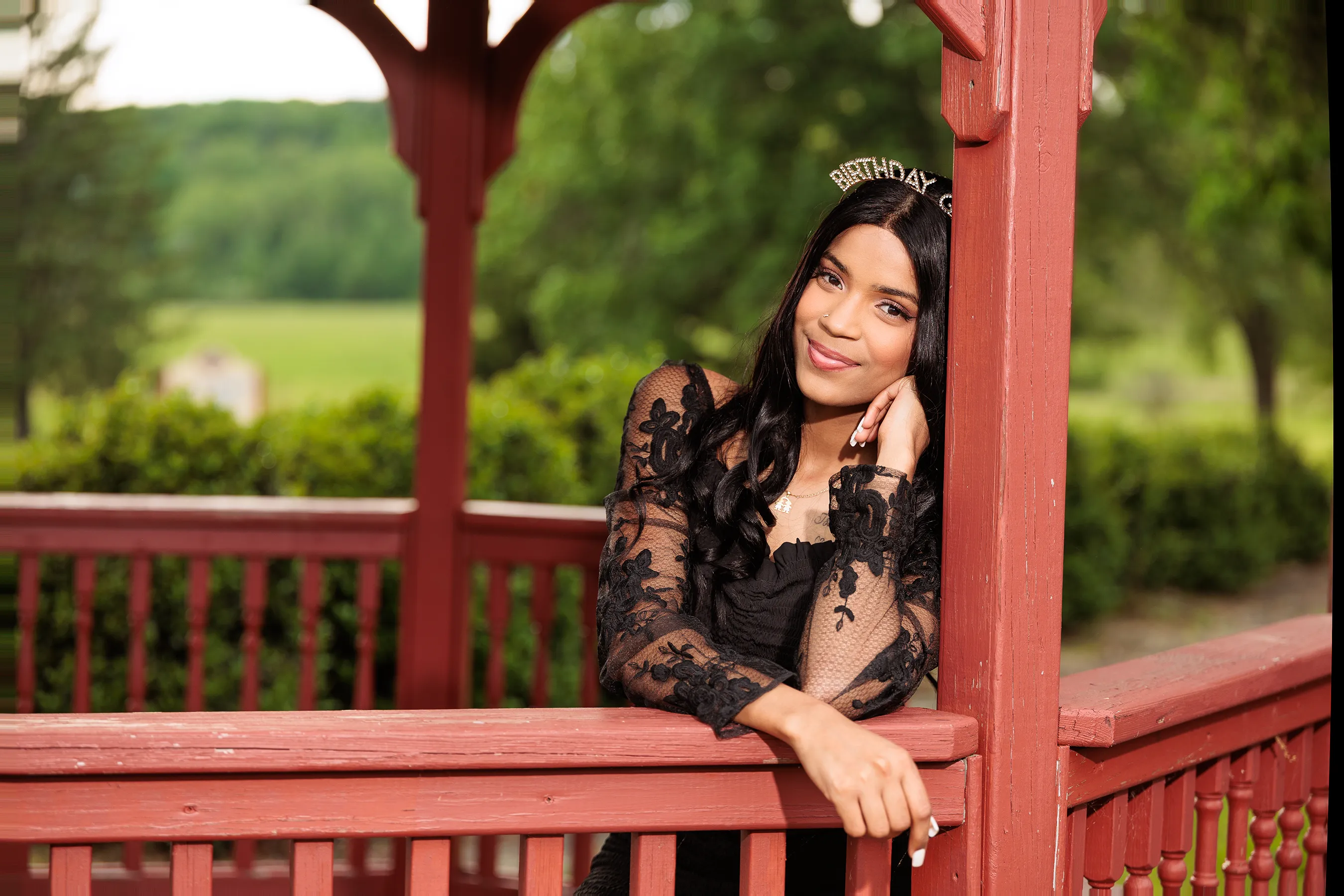 Birthday portrait of a young woman in a black lace dress posing inside a wooden gazebo at a park in Middletown NY, smiling softly during her outdoor photo session.