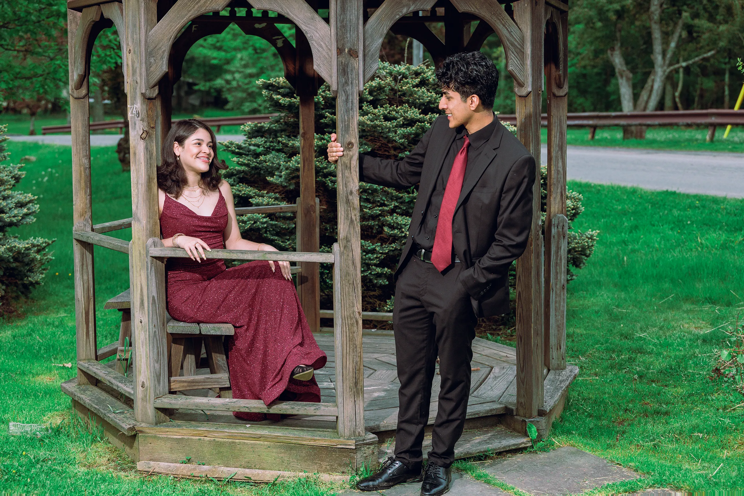 Prom portrait of two teens in coordinated burgundy and black formal outfits at De Hoyas Park in Monticello NY, smiling and posing together by a wooden gazebo during an outdoor session.
