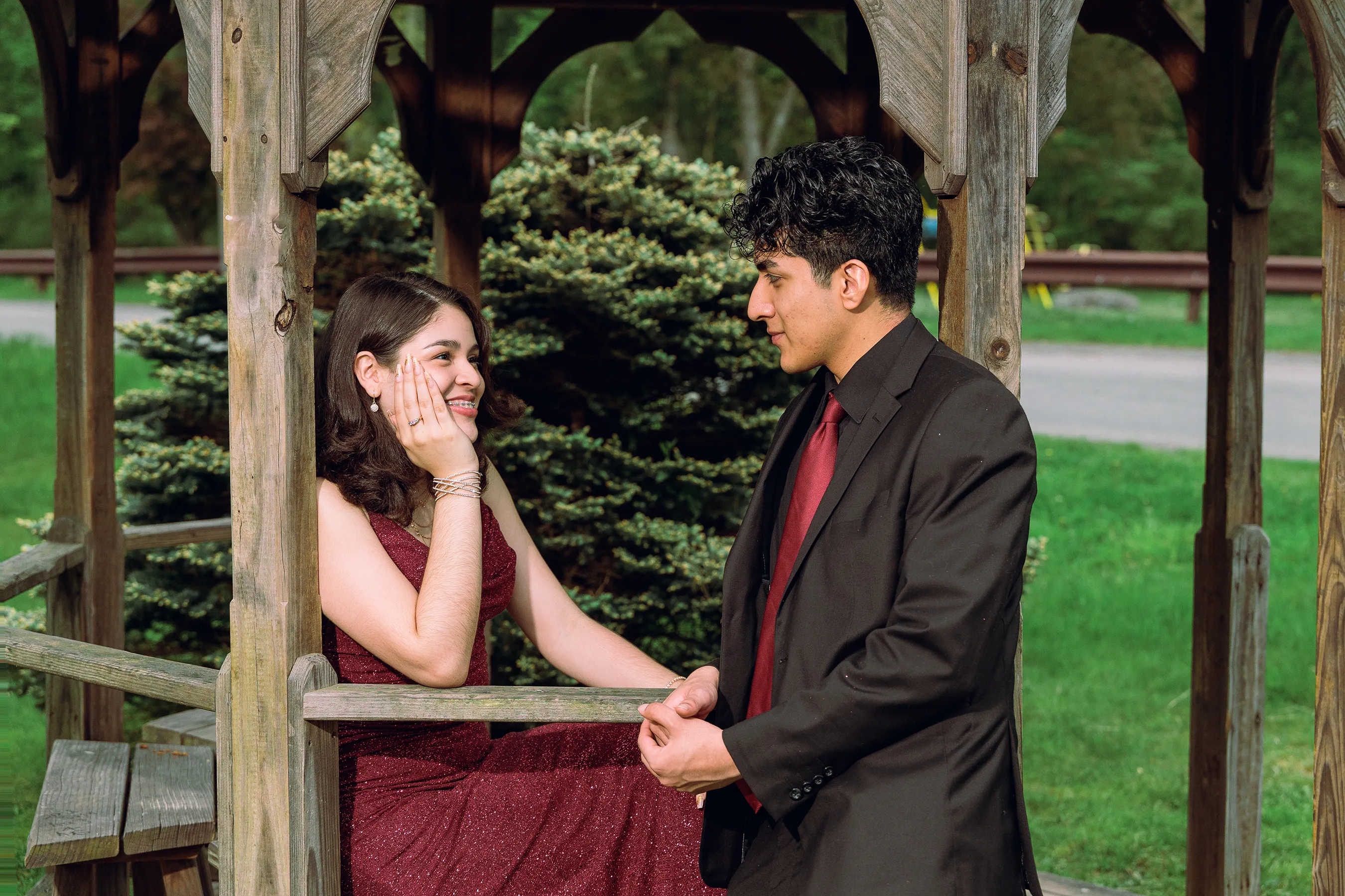 Prom portrait of two teens interacting naturally inside a wooden gazebo at De Hoyas Park in Monticello NY, dressed in coordinated burgundy and black outfits during an outdoor session.