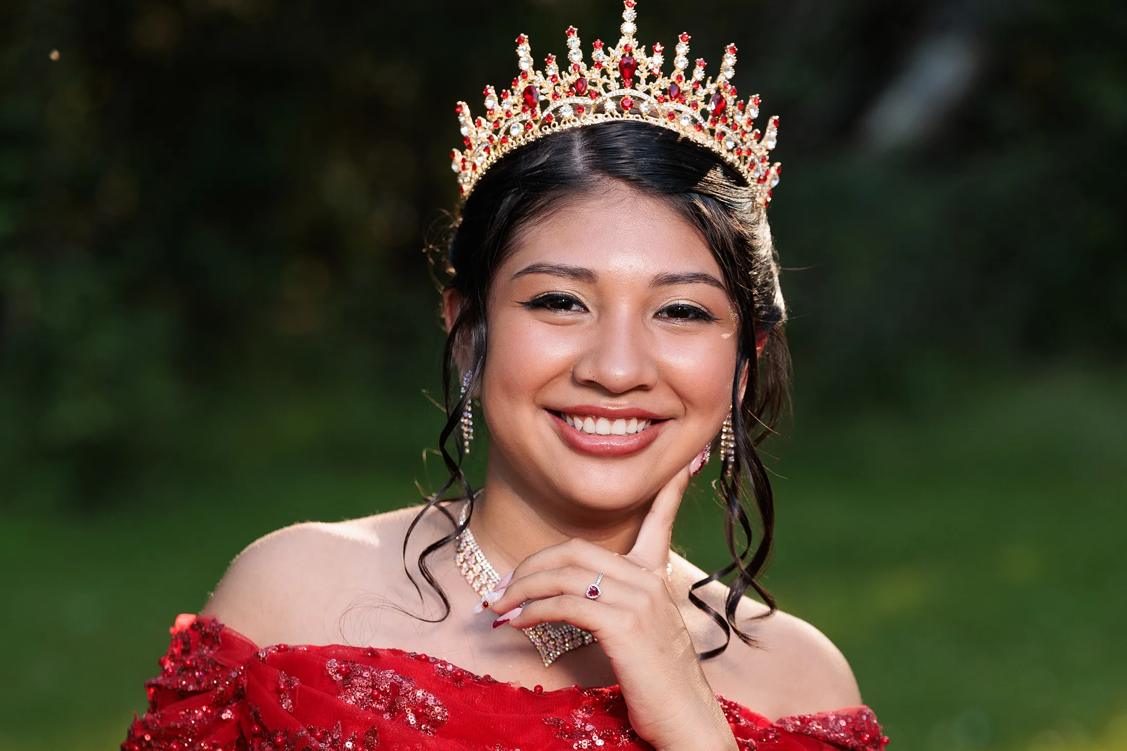Close-up quinceañera portrait taken in Glen Spey NY, young woman in a red gown and jeweled crown smiling with her hand on her chin under natural outdoor lighting. / Retrato en primer plano de una quinceañera en Glen Spey NY, joven con vestido rojo y corona con gemas, sonriendo con la mano en el mentón bajo luz natural.