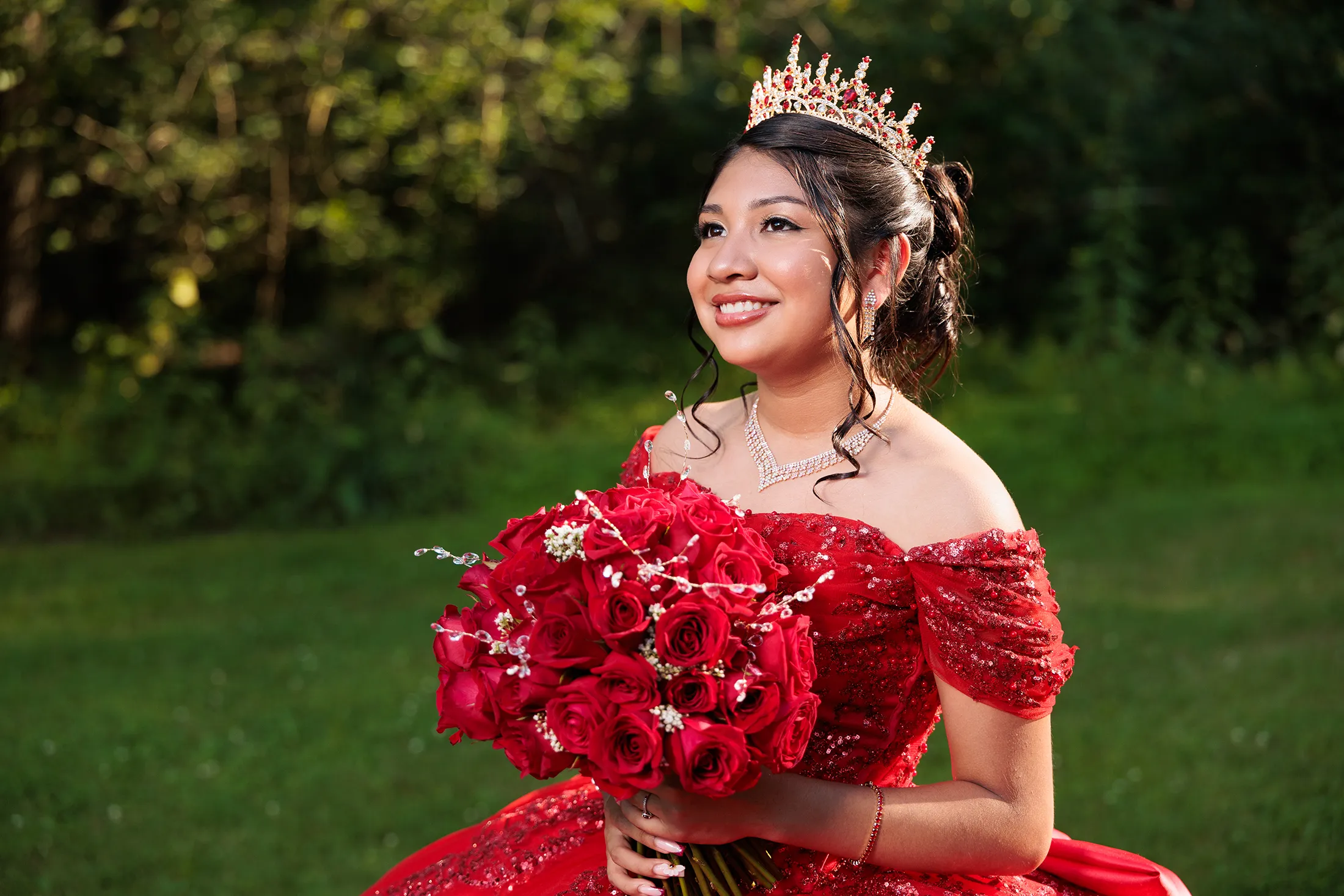 Outdoor quinceañera portrait in Glen Spey NY featuring a young woman in a red ballgown holding a bouquet of red roses, smiling with warm sunlight illuminating her face. / Retrato al aire libre de una quinceañera en Glen Spey NY con un vestido rojo y ramo de rosas rojas, sonriendo mientras la luz del sol ilumina su rostro.