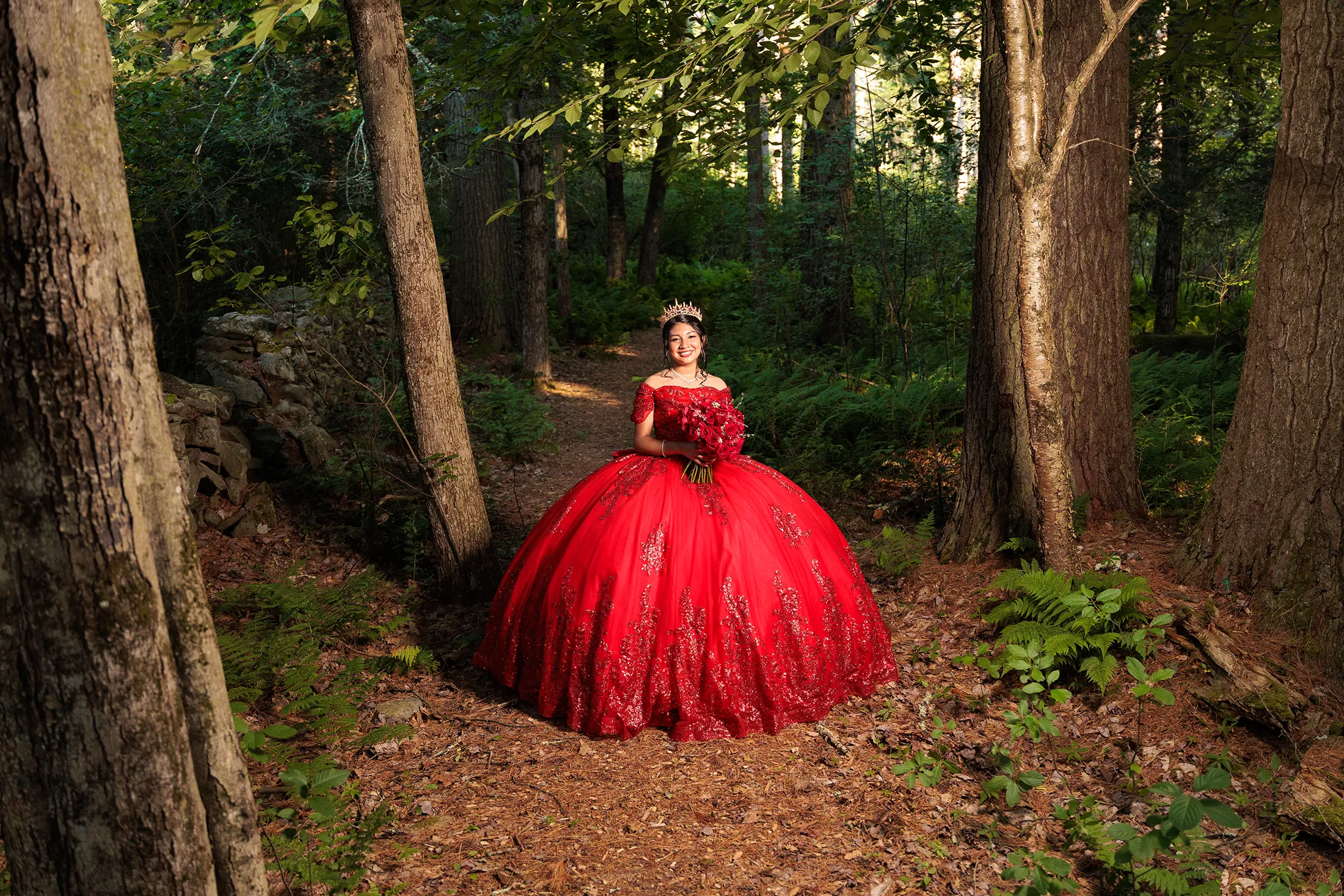 Full-body quinceañera portrait in Glen Spey NY showing a young woman in a bright red ballgown standing on a forest path, holding a rose bouquet with dramatic lighting highlighting the dress. / Retrato de cuerpo completo de una quinceañera en Glen Spey NY, luciendo un vestido rojo brillante sobre un sendero del bosque, con iluminación dramática que resalta el vestido.
