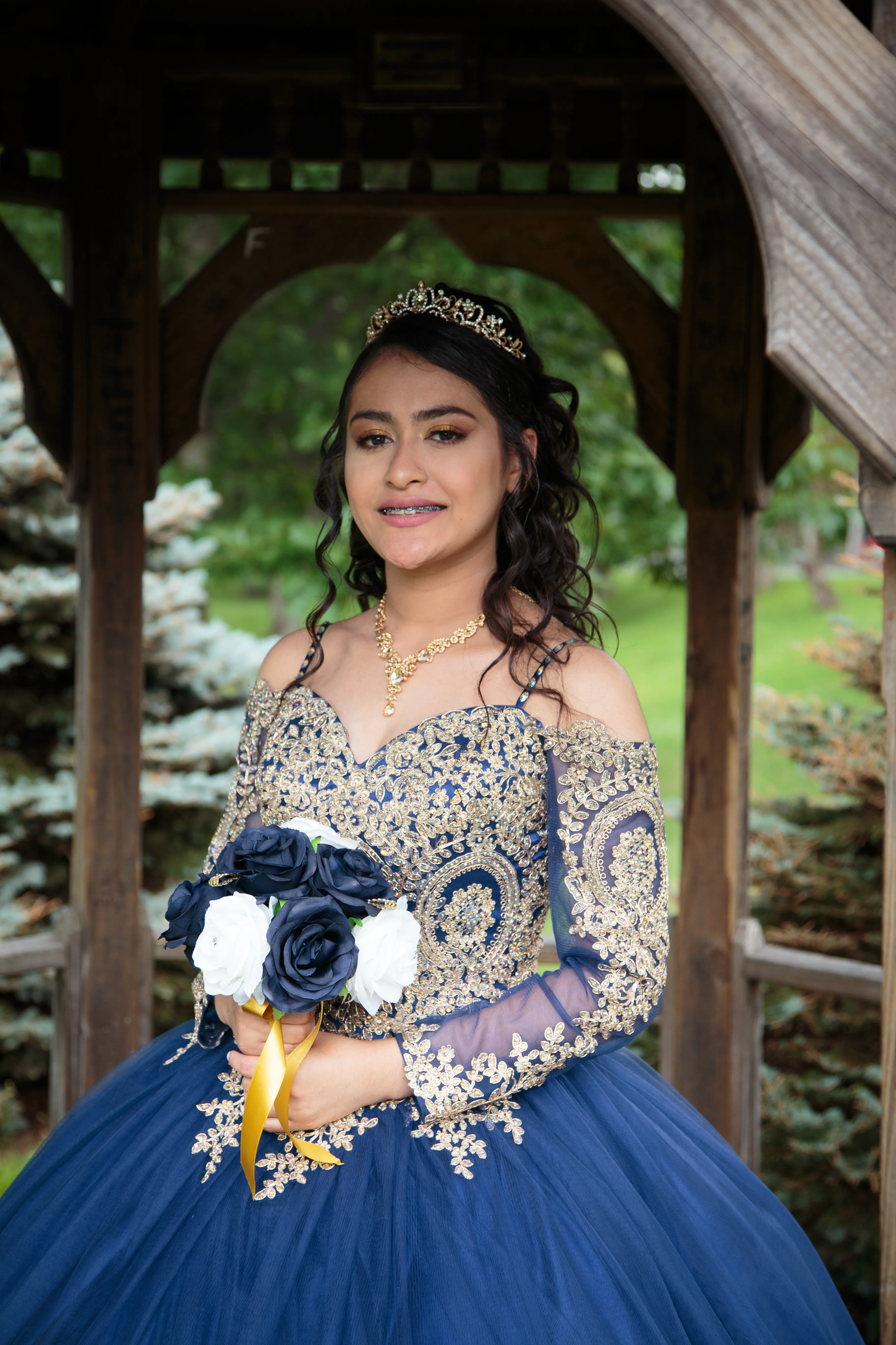 Quinceañera portrait taken at De Hoyas in Monticello NY — young girl in navy blue and gold quince dress holding bouquet, elegant outdoor Sweet 15 fotografía quinceañera bajo un gazebo de madera.