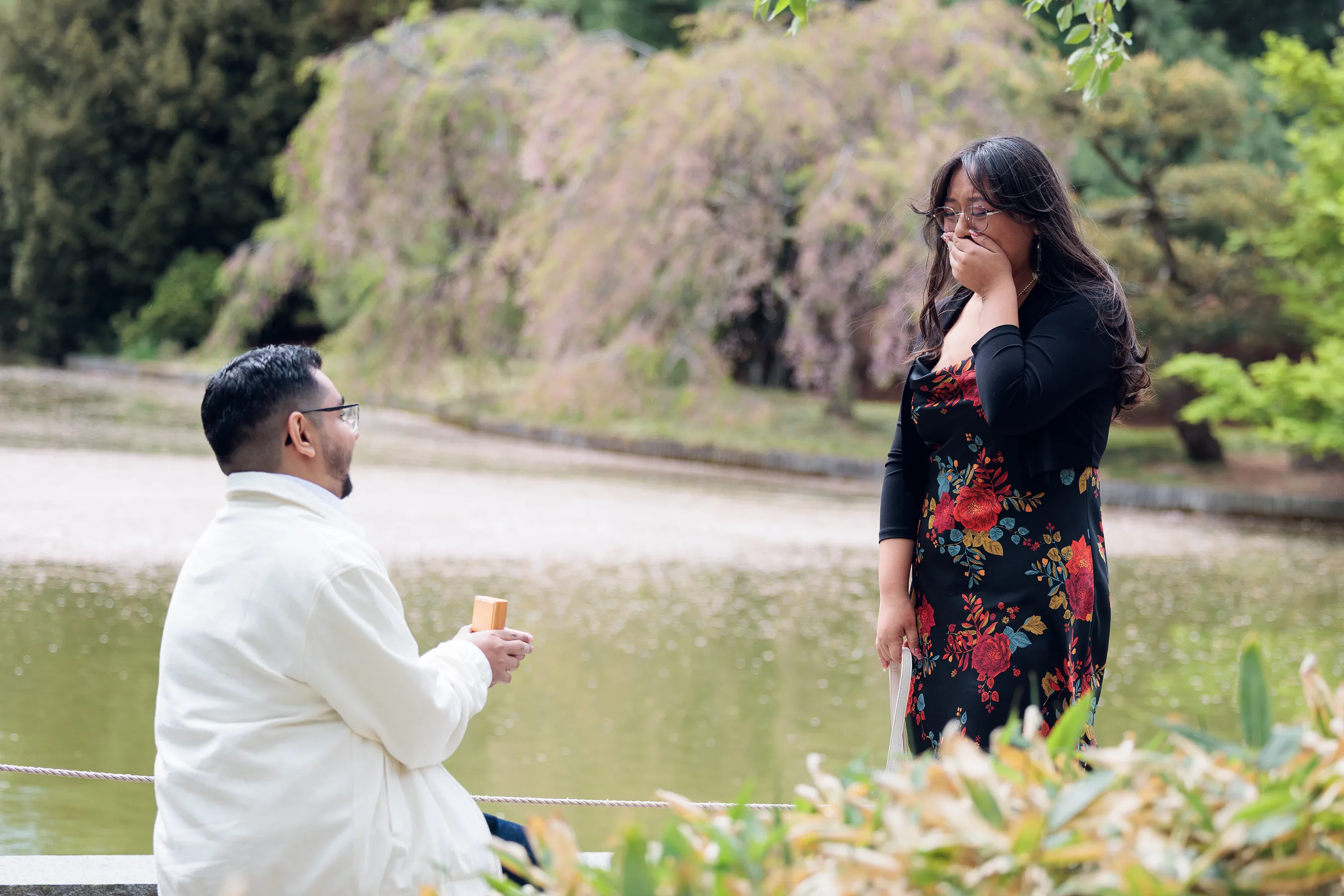 Emotional proposal moment at the Brooklyn Botanic Garden, with a man kneeling and presenting a ring while his partner reacts with surprise and joy by the water surrounded by spring greenery.